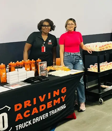 Two women standing by Driving Academy event table
